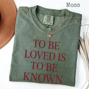 May include: A moss green t-shirt with the phrase "TO BE LOVED IS TO BE KNOWN" printed in red. The shirt has a crew neck and short sleeves. A gold necklace with a pendant is draped across the shirt.