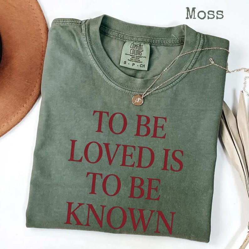 May include: A moss green t-shirt with the phrase "TO BE LOVED IS TO BE KNOWN" printed in red. The shirt has a crew neck and short sleeves. A gold necklace with a pendant is draped across the shirt.