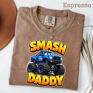 Może przedstawiać: Koszulka w kolorze espresso z grafiką niebieskiego monster trucka z napisem "SMASH DADDY" w kolorze ż&oacute;łtym i pomarańczowym. Koszulka ma r&oacute;wnież napis "BEST FATHER EVER" na ciężar&oacute;wce. Srebrny naszyjnik z wisiorkiem jest na koszulce.