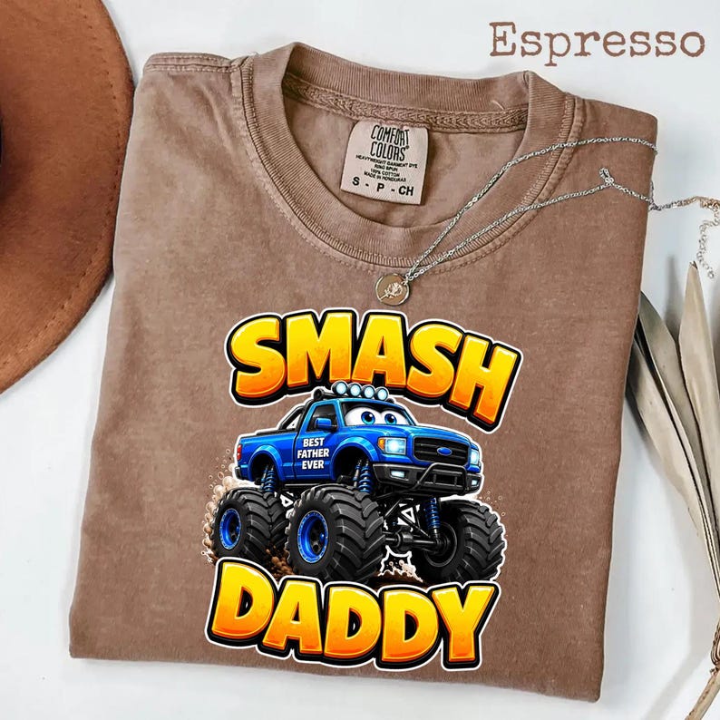 Może przedstawiać: Koszulka w kolorze espresso z grafiką niebieskiego monster trucka z napisem "SMASH DADDY" w kolorze ż&oacute;łtym i pomarańczowym. Koszulka ma r&oacute;wnież napis "BEST FATHER EVER" na ciężar&oacute;wce. Srebrny naszyjnik z wisiorkiem jest na koszulce.