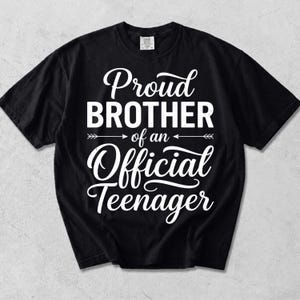 May include: A black short-sleeved t-shirt featuring white text that reads Proud BROTHER of an Official Teenager, with decorative arrows flanking the phrase of an. The design is centered on the chest of the garment, which has a crew neckline.