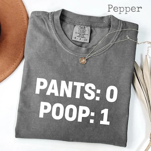 Könnte beinhalten: Ein graues Comfort Colors T-Shirt mit dem weißen Text "PANTS: 0 POOP: 1". Das Shirt hat einen Rundhalsausschnitt und kurze Ärmel. Eine silberne Halskette mit Anhänger befindet sich auf dem Shirt. Das Wort "Pepper" steht über dem Shirt.