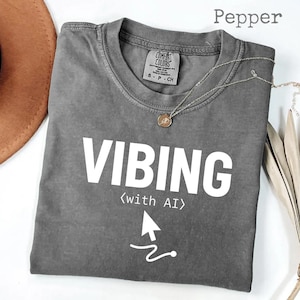 以下が含まれることがあります： 大きな白い文字で「VIBING」と書かれたグレーのComfort Colors Tシャツ。 下には矢印のグラフィックとともに「(with AI)」と書かれています。 ペンダント付きのシルバーのネックレスがシャツの上にかけられています。