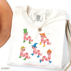 Op de afbeelding: Ivoor kleurig t-shirt met een grillig ontwerp van cartoonwormen met verschillende hoeden en laarzen. De tekst "IF A WORM WORE BOOTS, HOW WOULD IT WEAR THEM?" staat onderaan. Een gouden ketting ligt over het shirt.
