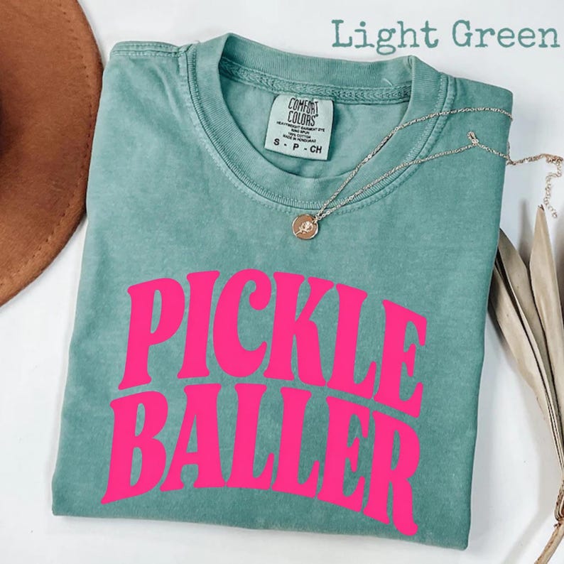 Op de afbeelding: Lichtgroen t-shirt met de tekst "PICKLE BALLER" in felroze. Het shirt heeft een ronde hals en korte mouwen. Een zilveren ketting met een kleine hanger is over het shirt gedrapeerd.