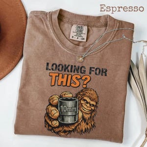 Könnte beinhalten: Espressofarbenes T-Shirt mit dem Text "LOOKING FOR THIS?" und einer Grafik eines lächelnden Bigfoot, der eine 10-mm-Stecknuss hält. Das Shirt hat einen Rundhalsausschnitt und kurze Ärmel. Eine silberne Halskette mit Anhänger ist ebenfalls sichtbar.
