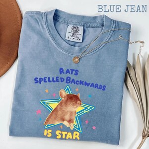 May include: Blue jean Comfort Colors t-shirt with the text "RATS SPELLED BACKWARDS IS STAR" and a graphic of a brown rat inside a yellow star. A gold necklace with a pendant is draped across the shirt.