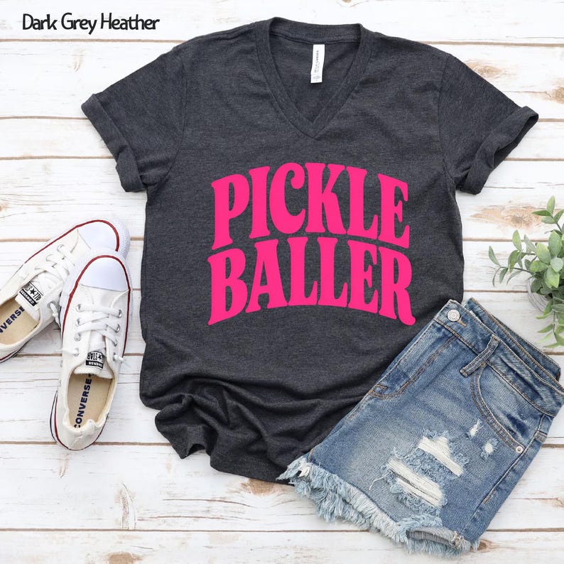 Op de afbeelding: Donkergrijze heather V-hals T-shirt met de tekst "PICKLE BALLER" in felroze. Het shirt wordt gecombineerd met een spijkerbroek en witte sneakers. De afbeelding is gemaakt op een licht houten oppervlak.
