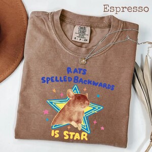 May include: Espresso-colored t-shirt with the text "RATS SPELLED BACKWARDS IS STAR" in blue and yellow. The graphic features a rat inside a star. A silver necklace with a pendant is draped over the shirt.