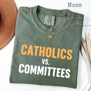 Op de afbeelding: Een mosgroen t-shirt met de woorden "CATHOLICS" in oranje en "VS. COMMITTEES" in wit. Het shirt heeft een ronde hals en korte mouwen. Een gouden ketting met een hanger is over het shirt gedrapeerd.