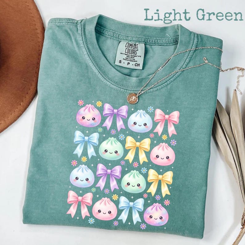 May include: A light green t-shirt featuring a repeating graphic pattern of smiling, round, pastel-colored dumplings interspersed with various bows in shades of pink, purple, blue, and yellow, accented by small, colorful flower icons.