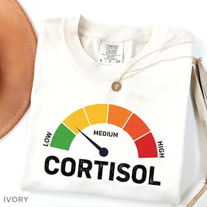 May include: Ivory Comfort Colors t-shirt featuring a gauge graphic with the word "CORTISOL" below. The gauge ranges from green to red, indicating levels. A gold necklace is draped across the shirt. The shirt is ivory in colour.