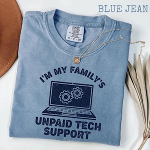 Op de afbeelding: Blauw Comfort Colors T-shirt in jeanskleur met de tekst "I'M MY FAMILY'S UNPAID TECH SUPPORT" en een laptopafbeelding. Een gouden ketting met een hanger is over het shirt gedrapeerd. Het shirt is op een wit oppervlak gevouwen.