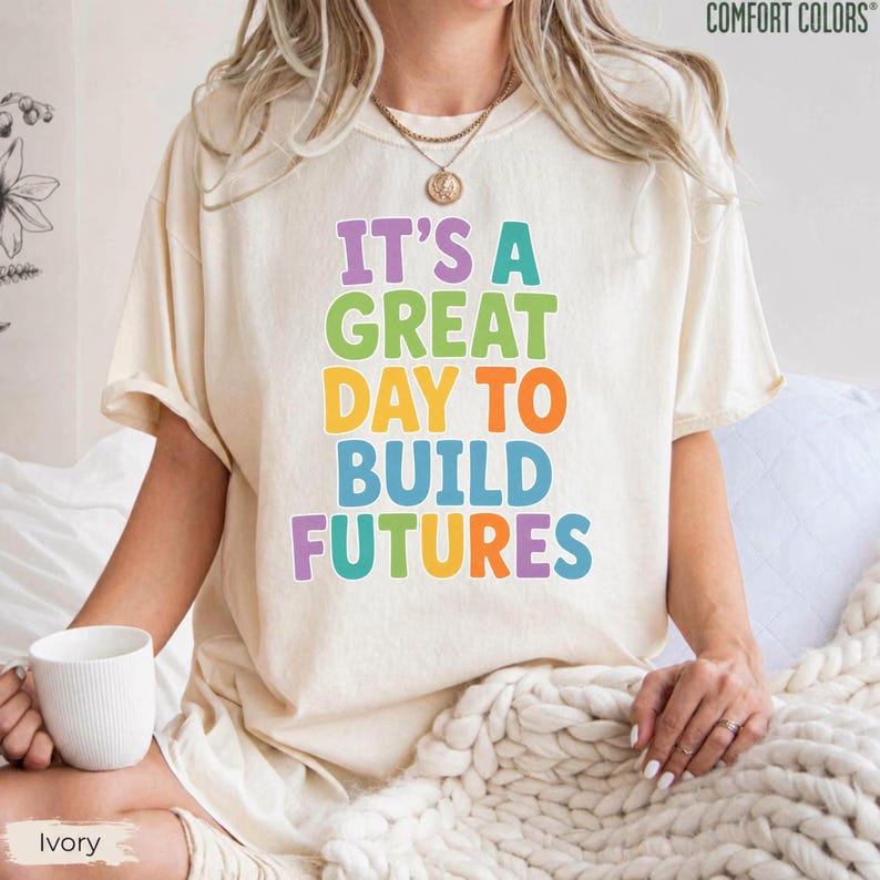 Puede incluir: Camiseta de color marfil con el texto "IT'S A GREAT DAY TO BUILD FUTURES" en letras de colores. La camiseta es de la marca Comfort Colors y tiene un corte relajado.