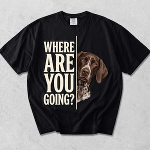 Puede incluir: Una camiseta negra de manga corta con el texto en color crema WHERE ARE YOU GOING? impreso verticalmente en el lado izquierdo, junto a una ilustración realista de un perro marrón y blanco asomándose por el lado derecho de la camiseta.