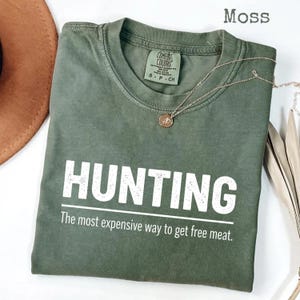 May include: A moss green t-shirt with the word "HUNTING" in large white letters. Below, the text reads, "The most expensive way to get free meat." A gold necklace with a pendant is draped across the shirt.