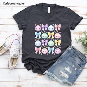 May include: A dark grey heather V-neck t-shirt featuring a repeating pattern of pastel-colored kawaii-style dumplings and bows. The graphic includes pink, blue, purple, and yellow elements arranged in a grid, with small flower accents throughout.