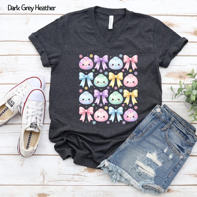 May include: A dark grey heather V-neck t-shirt featuring a repeating pattern of pastel-colored kawaii-style dumplings and bows. The graphic includes pink, blue, purple, and yellow elements arranged in a grid, with small flower accents throughout.