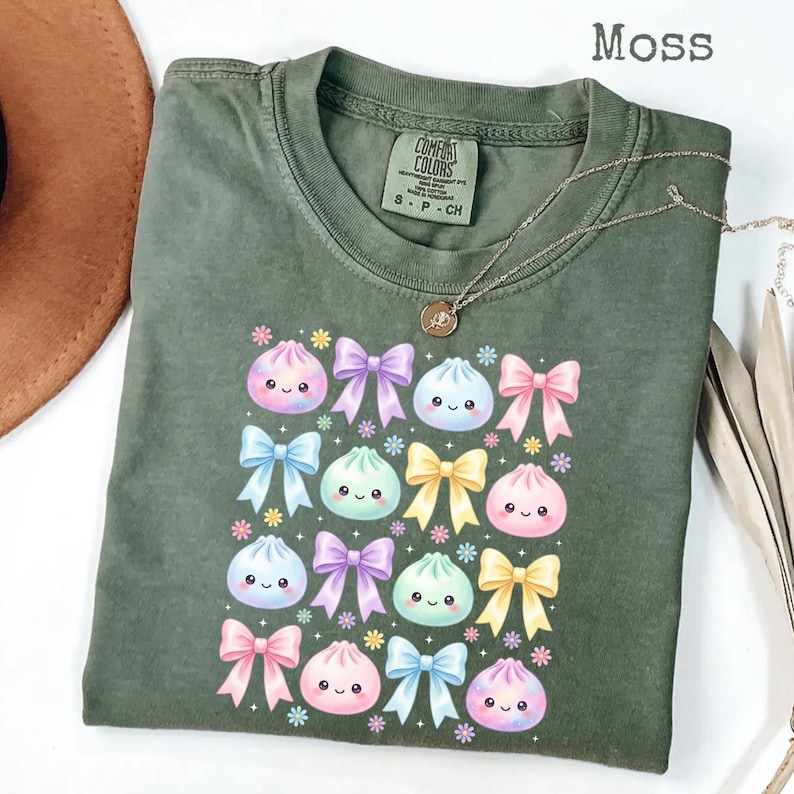 May include: A moss green crew neck t-shirt featuring a graphic print of various pastel-colored bows and smiling dumpling characters. The shirt has a Comfort Colors brand label at the inner neckline and is paired with a delicate gold-tone necklace.