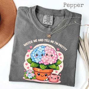 Puede incluir: Camiseta gris con el texto "WATER ME AND TELL ME I'M PRETTY" y una ilustración de dibujos animados de flores azules y rosas en una maceta. La camiseta tiene cuello redondo y mangas cortas. Un collar plateado está sobre la camiseta.