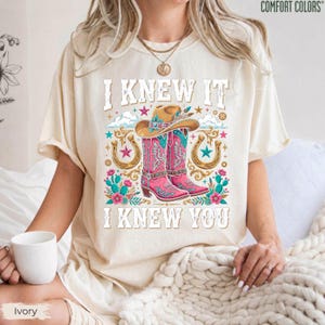 May include: An ivory short-sleeve t-shirt featuring a graphic print of a cowboy hat and pink cowboy boots surrounded by horseshoes, stars, and cacti. The text reads I KNEW IT I KNEW YOU. The shirt has a crew neckline and a relaxed fit.