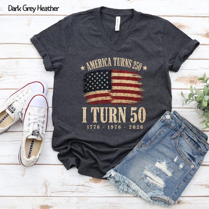 May include: Dark grey heather V-neck t-shirt with the text "America Turns 250, I Turn 50" and an American flag design. Includes white Converse sneakers and denim shorts. The shirt's design features the years 1776, 1976, and 2026.