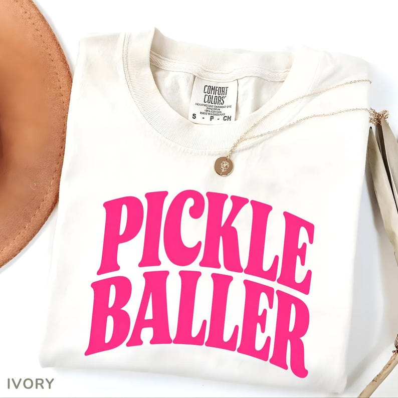 Op de afbeelding: Ivoor kleurig t-shirt met de woorden "PICKLE BALLER" in felroze, een gouden ketting en een bruine hoed. Het shirt heeft een ronde hals en een label met de tekst "COMFORT COLORS". Het shirt is gemaakt van zwaar geverfd katoen.