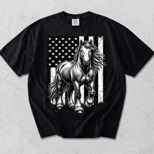 以下が含まれることがあります： 黒の半袖Tシャツで、かすれたアメリカ国旗のデザインの前に立つ馬のグラフィックプリントが施されています。馬は黒、白、グレーの色調で描かれており、流れるようなたてがみと詳細な筋肉の質感が表現されています。