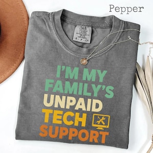 Może przedstawiać: Szara koszulka Comfort Colors z napisem "I'M MY FAMILY'S UNPAID TECH SUPPORT" w kolorze zielonym, żółtym i pomarańczowym. Projekt zawiera ikonę monitora komputera z kluczem i śrubokrętem. Srebrny naszyjnik z wisiorkiem jest ułożony na koszulce.