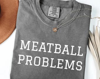 May include: A gray Comfort Colors t-shirt with the words "MEATBALL PROBLEMS" in white block letters. A silver necklace with a small pendant rests on the shirt. The shirt is folded, and a brown hat is visible in the background.
