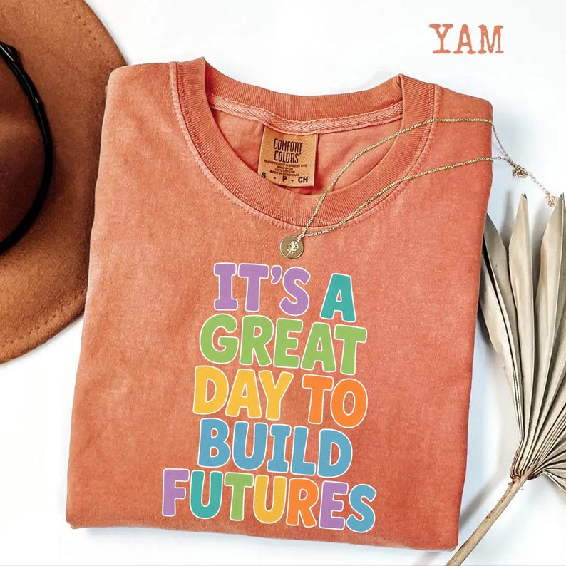 Puede incluir: Una camiseta de color coral con el texto "IT'S A GREAT DAY TO BUILD FUTURES" en letras de colores. La camiseta tiene cuello redondo y mangas cortas. Tambi&eacute;n se ven un collar dorado y un sombrero marr&oacute;n.
