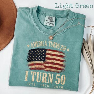 May include: Light green t-shirt with the text "AMERICA TURNS 250" and "I TURN 50" with the years 1776-1976-2026. The shirt features a distressed American flag graphic. A silver necklace with a pendant is on the shirt.