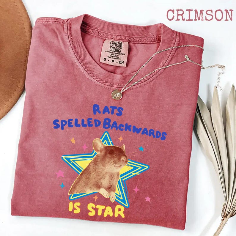 May include: Crimson-colored t-shirt with the text "RATS SPELLED BACKWARDS IS STAR" and a graphic of a rat inside a star. A silver necklace with a pendant is draped across the shirt.