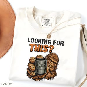Könnte beinhalten: Elfenbeinfarbenes T-Shirt mit dem Text "LOOKING FOR THIS?" in Schwarz und Orange. Eine Cartoon-Bigfoot-Figur hält einen 10-mm-Steckschlüssel. Das Shirt hat ein Comfort Colors-Etikett und eine goldene Halskette.