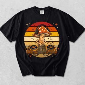 Könnte beinhalten: Ein schwarzes T-Shirt mit einer Grafik eines lächelnden Pilzes, der Kopfhörer trägt und an einem DJ-Pult auflegt. Das Motiv befindet sich vor einem kreisförmigen Hintergrund mit horizontalen Streifen in Orange-, Gelb- und Rottönen.