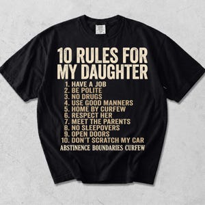 May include: A black short-sleeved t-shirt featuring the beige text 10 RULES FOR MY DAUGHTER followed by a numbered list of ten rules including Have a Job, Be Polite, No Drugs, Use Good Manners, Home by Curfew, Respect Her, Meet the Parents, No Sleepovers, Open Doors, Don't Scratch My Car, and the words Abstinence Boundaries Curfew.