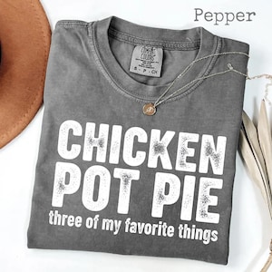 Pode incluir: Uma camiseta cinza Comfort Colors com o texto branco "CHICKEN POT PIE three of my favorite things". A camiseta tem gola redonda e mangas curtas. Um colar de prata com um pingente está sobre a camiseta.