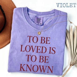 May include: A violet-colored t-shirt with the text "TO BE LOVED IS TO BE KNOWN" in a bold, dark red font. A gold necklace with a pendant rests on the shirt. The shirt has a crew neck and a small tag.