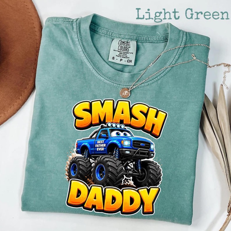 Może przedstawiać: Jasnozielona koszulka z grafiką niebieskiego monster trucka z napisem "SMASH DADDY" w kolorze ż&oacute;łtym i pomarańczowym. Na ciężar&oacute;wce widnieje napis "BEST FATHER EVER". Srebrny naszyjnik jest zarzucony na koszulkę.