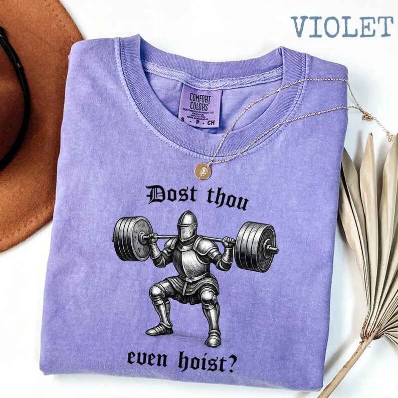 May include: Violet-colored t-shirt with a graphic of a knight lifting weights, and the text "Dost thou even hoist?" A gold necklace is draped across the shirt. The shirt has a crew neck and short sleeves.