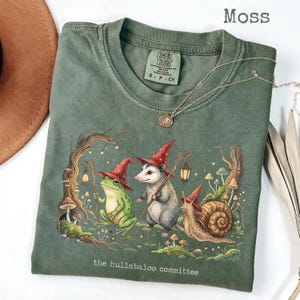 Op de afbeelding: Een mosgroen t-shirt met een afbeelding van een kikker, een opossum en een slak, die allemaal rode puntmutsen dragen. De dieren bevinden zich in een bos met paddenstoelen en lantaarns. De tekst "the hullabaloo committee" staat onderaan gedrukt.