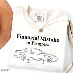 Könnte beinhalten: Elfenbeinfarbenes T-Shirt mit dem Text "Financial Mistake in Progress" über einer Strichzeichnung eines Autos auf Wagenhebern. Das Shirt hat ein Comfort Colors-Etikett und eine goldene Halskette.