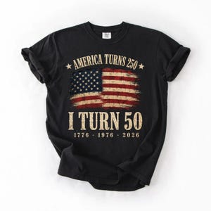 May include: Black t-shirt with a distressed American flag graphic. The text reads "America Turns 250" above the flag and "I Turn 50" below, with the years 1776-1976-2026. A patriotic design.