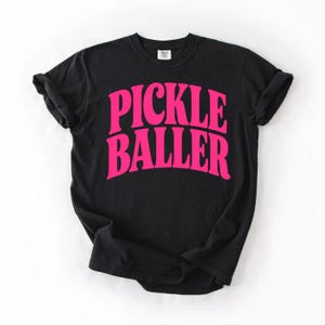 Op de afbeelding: Zwart T-shirt met de tekst "PICKLE BALLER" in felroze, vette, gebogen letters. Het shirt heeft een ronde hals, korte mouwen en een relaxte pasvorm. De tekst is gecentreerd op de voorkant.