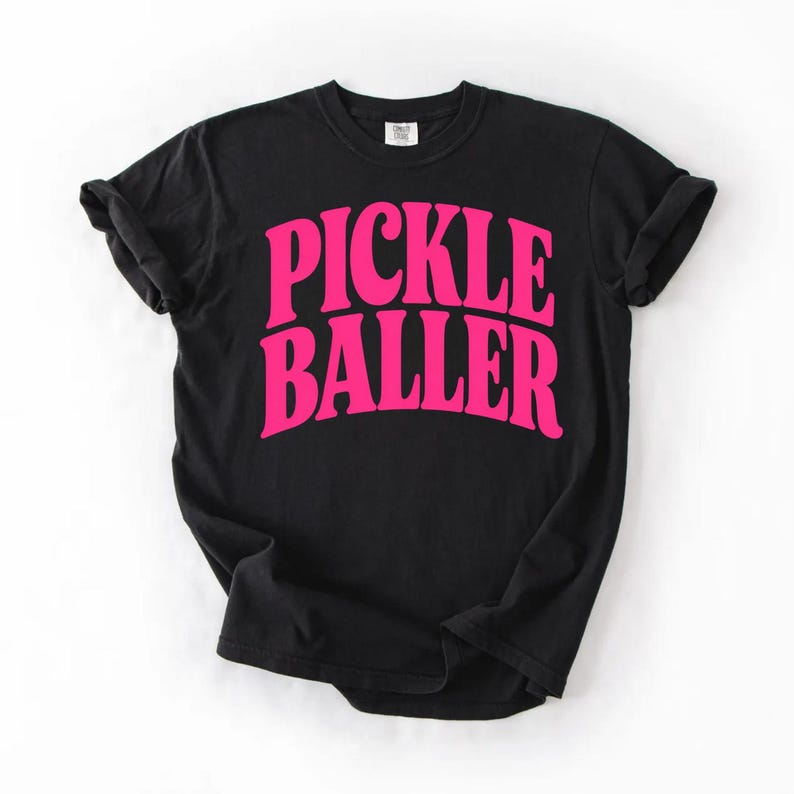 Op de afbeelding: Zwart T-shirt met de tekst "PICKLE BALLER" in felroze, vette, gebogen letters. Het shirt heeft een ronde hals, korte mouwen en een relaxte pasvorm. De tekst is gecentreerd op de voorkant.