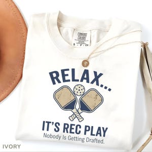 Könnte beinhalten: Cremefarbenes T-Shirt mit dem Text "RELAX... IT'S REC PLAY" in Marineblau, zusammen mit einer Pickleball-Grafik. Das Shirt hat auch den Text "Nobody Is Getting Drafted." Eine goldene Halskette und ein Hut sind ebenfalls sichtbar.
