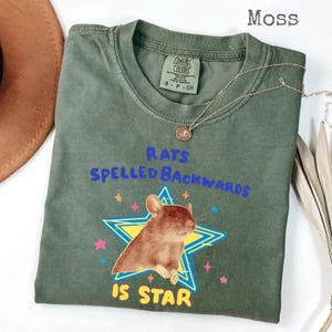 May include: Moss green t-shirt with a graphic of a brown rat inside a yellow and blue star. The text reads "RATS SPELLED BACKWARDS IS STAR" in blue and yellow. A silver necklace with a pendant is draped across the shirt.