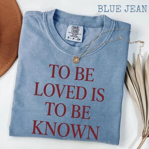 May include: A blue jean-colored t-shirt with the phrase "TO BE LOVED IS TO BE KNOWN" in red lettering. A gold necklace with a pendant rests on the shirt. The shirt has a Comfort Colors tag.