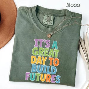 Puede incluir: Camiseta verde musgo con el texto "IT'S A GREAT DAY TO BUILD FUTURES" en letras coloridas. Un collar dorado con un colgante est&aacute; drapeado sobre la camiseta. La camiseta est&aacute; doblada y colocada sobre una superficie blanca.