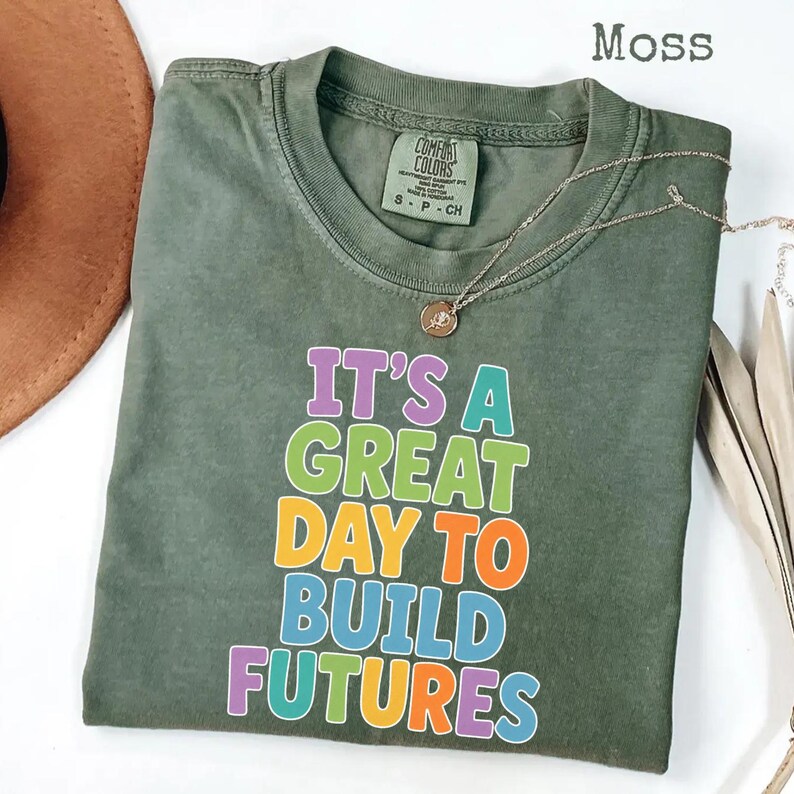 Puede incluir: Camiseta verde musgo con el texto "IT'S A GREAT DAY TO BUILD FUTURES" en letras coloridas. Un collar dorado con un colgante est&aacute; drapeado sobre la camiseta. La camiseta est&aacute; doblada y colocada sobre una superficie blanca.